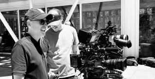 Behind-the-scenes shot of a videographer filming an actor in a classic car during daytime.